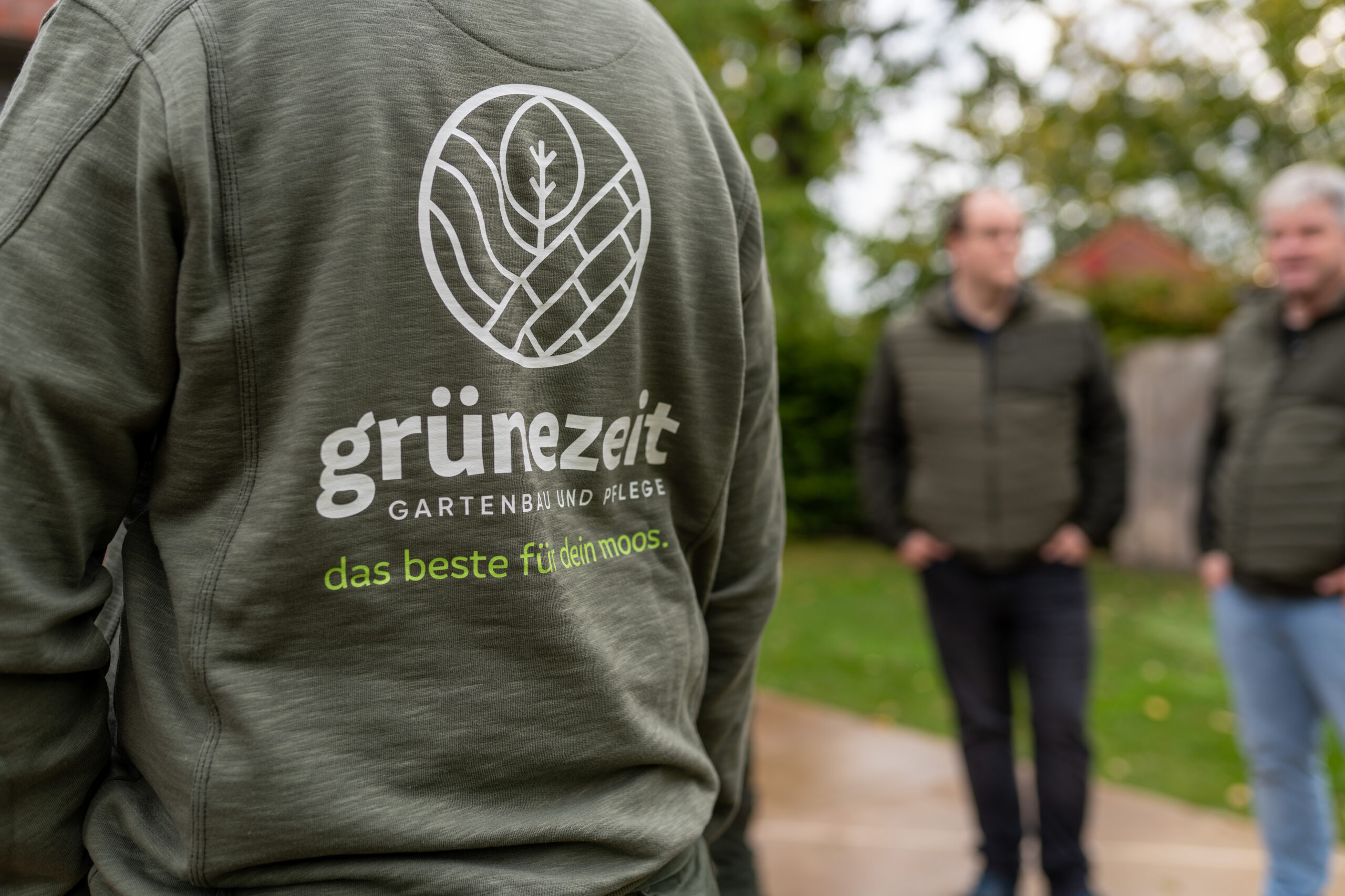 grünezeit-Logo auf der Arbeitskleidung mit Kollegen im Hintergrund